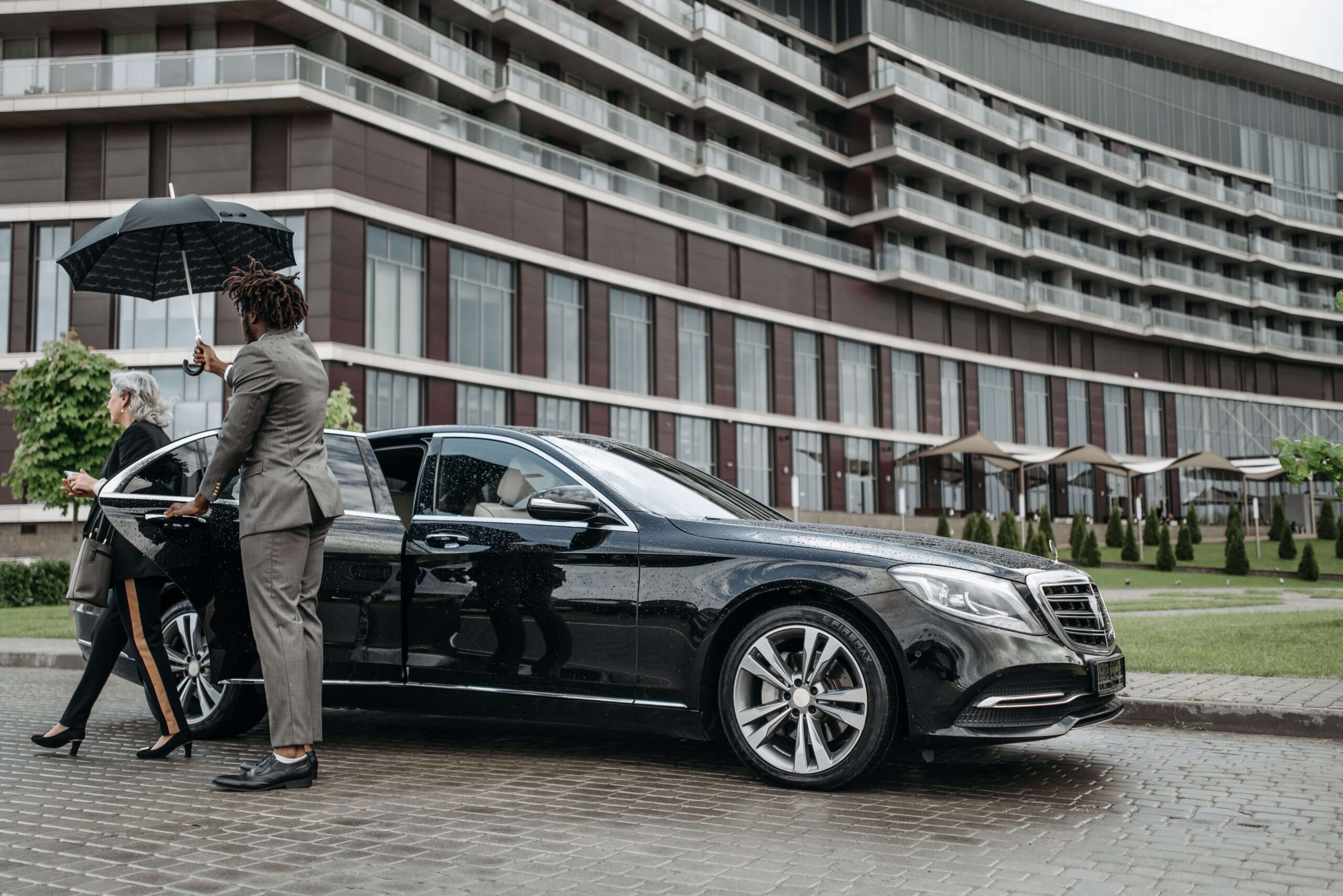 Chauffeur holding an umbrella for an elegant passenger entering a black Mercedes S-Class outside a hotel in Los Angeles